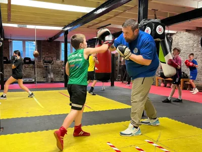 A boy in a green vest, black shorts  and red boots punches a hand pad being held by a bearded man in a blue t-shirt and cream trousers, with other young boxers in the background