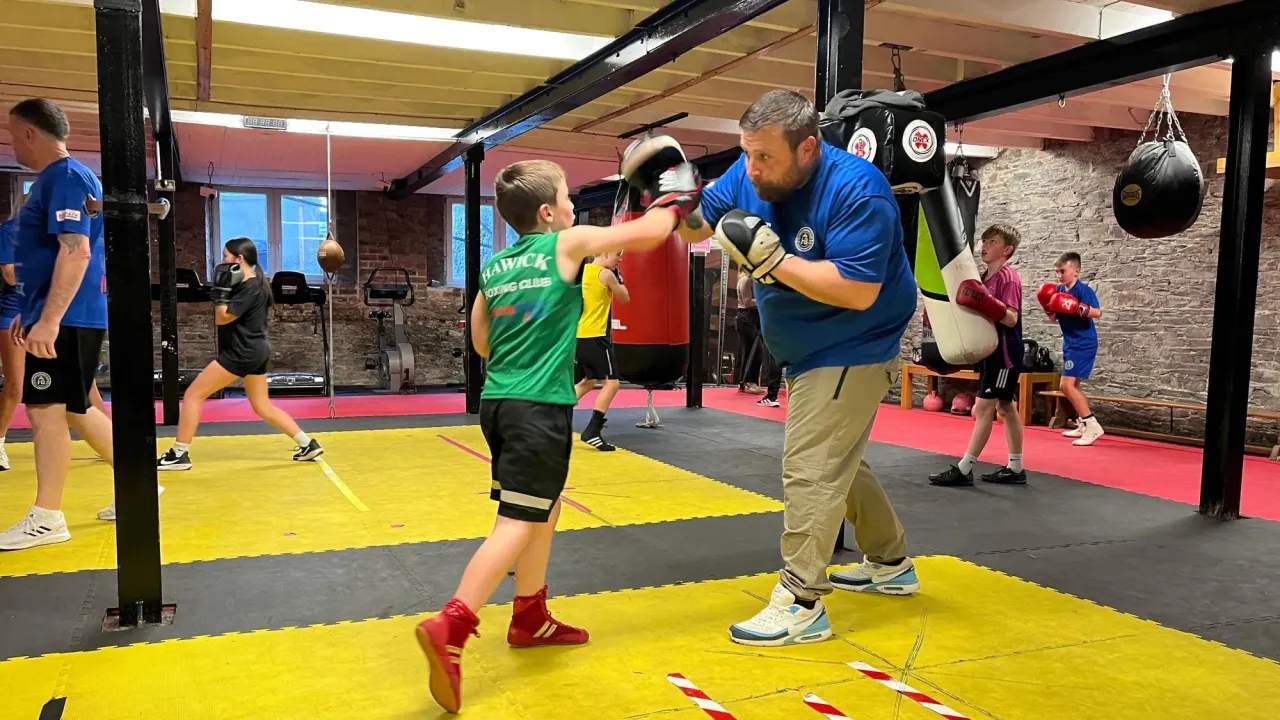 A boy in a green vest, black shorts and red boots punches a hand pad being held by a bearded man in a blue t-shirt and cream trousers, with other young boxers in the background