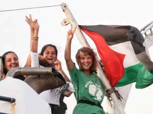 Greta Thunberg and other female crew members of the Global Sumud Flotilla are smiling and waving from a boat. A Palestinian flag which is tied to the vessel is flying in the breeze. Greta has short blonde hair and is wearing a green and white t-shirt 