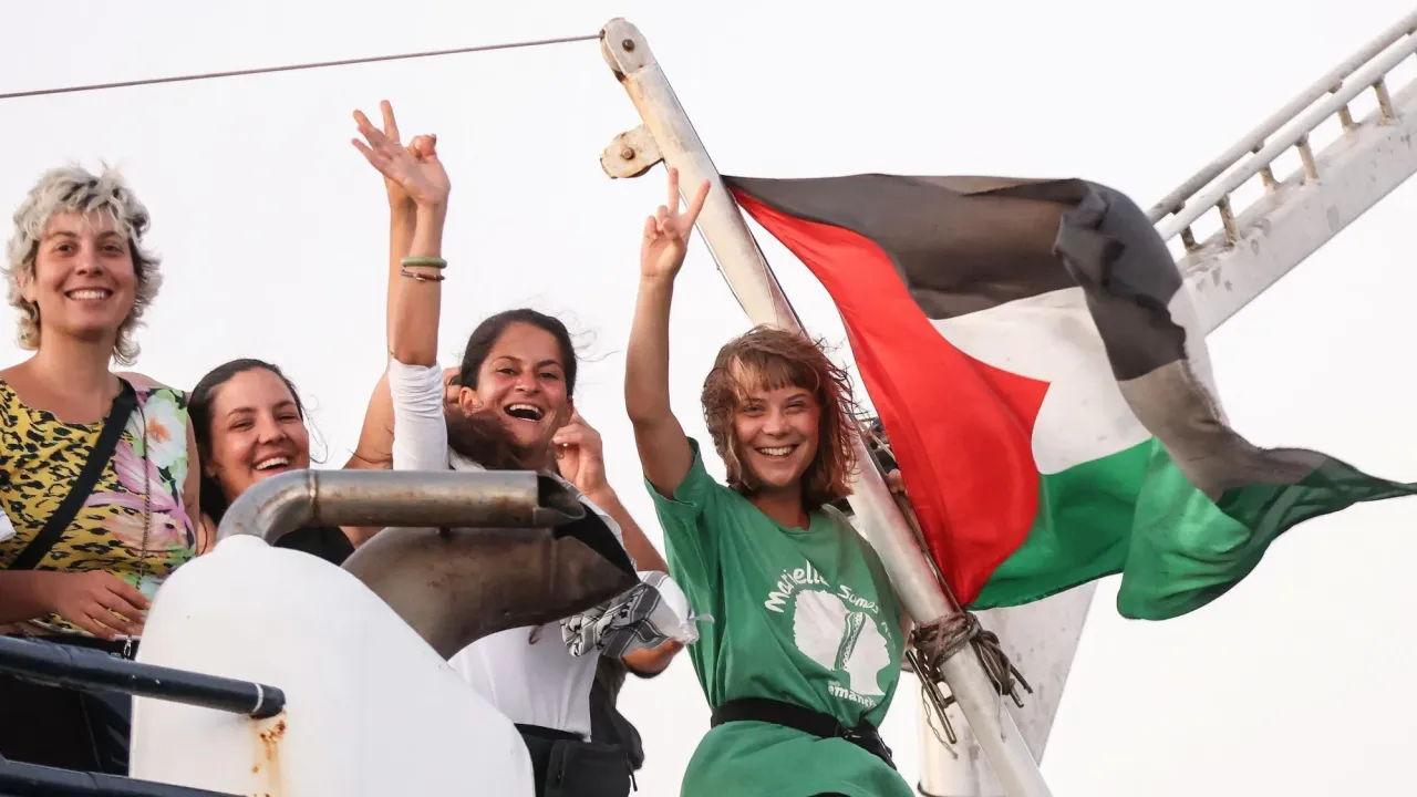 Greta Thunberg and other female crew members of the Global Sumud Flotilla are smiling and waving from a boat. A Palestinian flag which is tied to the vessel is flying in the breeze. Greta has short blonde hair and is wearing a green and white t-shirt 