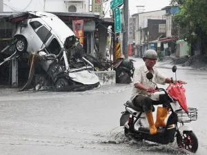 A man wearing a bike helmet, short-sleeved shirt and orange wellington boots rides his scooter past a damaged car that is upturned and leaning on the side of a building in Hualien county, Taiwan.