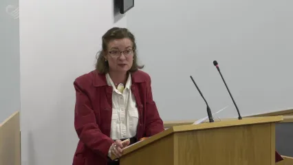 Eluned Morgan standing at a light wooden lectern in the temporary Senedd chamber responding to a question from Conservative group leader Darren Millar. She is wearing glasses, a white shirt and red jacket. Her right hand, in which she is holding a white coloured pen, is resting on the edge of the lectern.