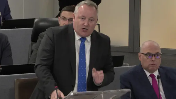 Darren Millar on his feet asking a question during First Minister's Questions in the temporary Senedd chamber in Cardiff Bay. He is wearing a blue tie with light spots, a light shirt and dark suit.