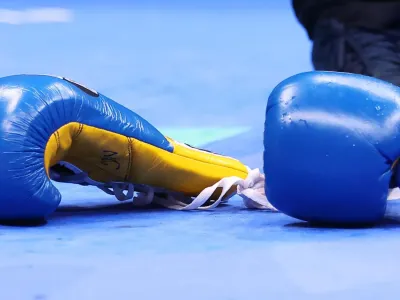 Two royal blue and yellow boxing gloves are seen on lying on a light blue canvas, with the navy blue left shoe and lower left leg of an official seen in the distance. The white laces of one glove can be seen hanging loose on the canvas