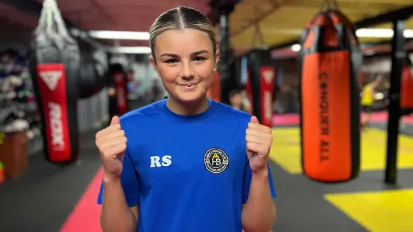 A girl with blonde-streaked hair, wearing a blue t-shirt, smiles at the camera. Behind her are punchbags