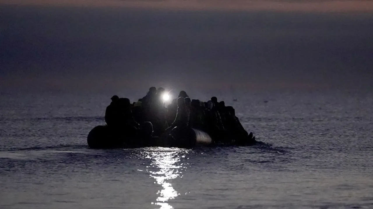 A silhouette of a migrant boat at twilight or sunrise, with a torch being shone, on water off the coast of Gravelines, France. The sky is tinged with red