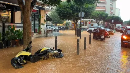 A street flooded with muddy water in Ibiza town features a yellow motorbike fallen on its side and some cars driving through the brown water.