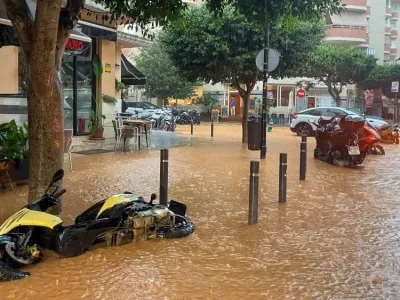 A street flooded with muddy water in Ibiza town features a yellow motorbike fallen on its side and some cars driving through the brown water.