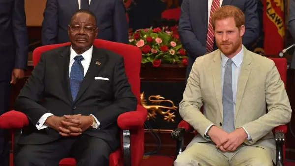 Peter Mutharika, wearing a black suit, sits next Prince Harry, who wears a beige suit. They are both seated in red chairs and figures stand behind them.