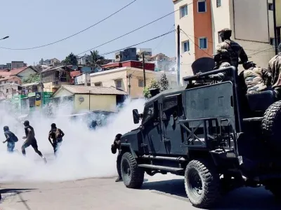 Riot Police in a heavily armoured bear-cat kind of vehicle drive behind three protesters who are running away through tear gas smoke.