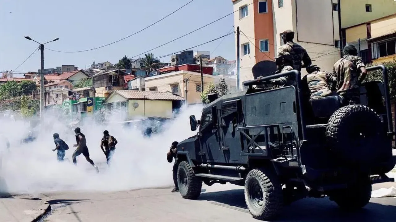 Riot Police in a heavily armoured bear-cat kind of vehicle drive behind three protesters who are running away through tear gas smoke.