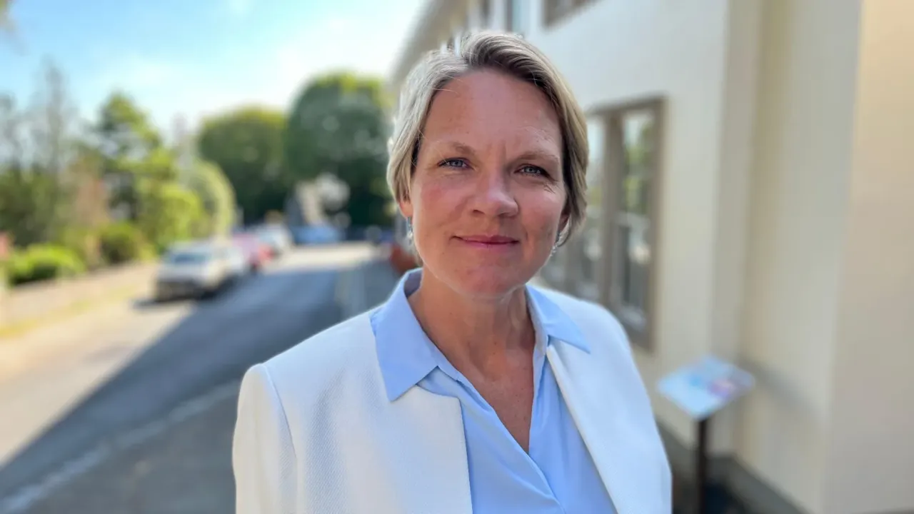 Woman in her 40s with short blonde hair. She wears a white blazer and a light blue shirt