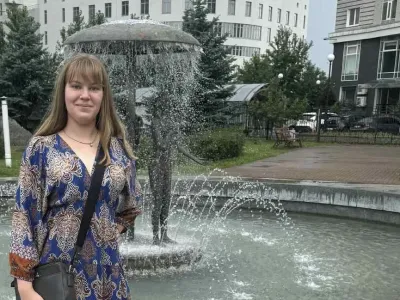 Sasha Shevchenko stands by a fountain which has a mushroom with figures underneath. She is wearing a blue dress and has a handbag over her shoulder. She has long blonde hair with a fringe and is wearing a necklace.