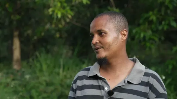 Peter Wambugu, with short hair, is wearing a gray and black striped polo shirt and standing outdoors in a lush, green environment with trees and foliage in the background."