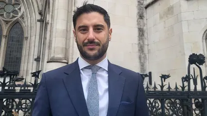 James Netto is in a suit standing in front of the Royal Courts of Justice, a historic Gothic-style building with large arched windows and ornate stonework. The building&#39;s name is visible on the wall behind him.