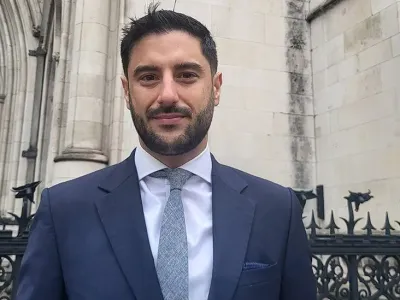 James Netto is in a suit standing in front of the Royal Courts of Justice, a historic Gothic-style building with large arched windows and ornate stonework. The building&#39;s name is visible on the wall behind him.