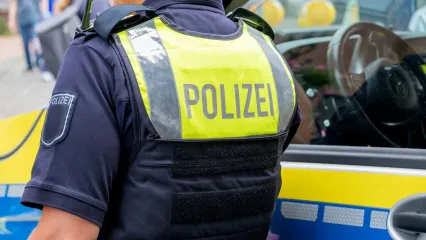 A German police officer&#39;s back features a fluorescent police vest with the words &quot;Polizei&quot; - police in German - on it. They are standing next to a police car.