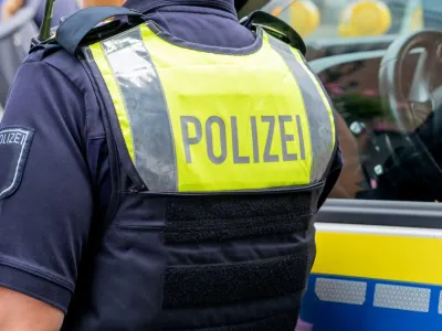 A German police officer&#39;s back features a fluorescent police vest with the words &quot;Polizei&quot; - police in German - on it. They are standing next to a police car.