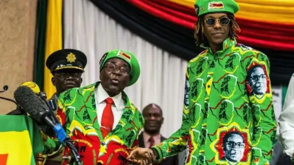 Former President Robert Mugabe standing to the left, next to his son, Robert Mugabe junior, who is standing to the right. The pair are shaking hands and are dressed in bright green suits that has the former presidents face on it. Both President Mugabe and his son, are wearing glasses, although his son is wearing dark sunglasses, while he is wearing what appears to be prescription glasses. Both men have bright green berets on that are emblazoned with the flag of Zimbabwe. 