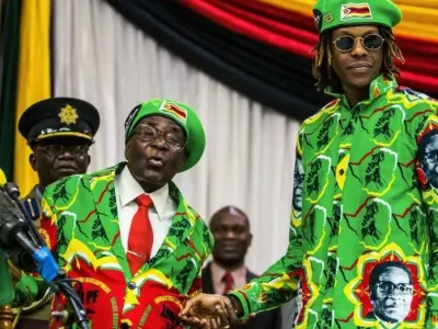 Former President Robert Mugabe standing to the left, next to his son, Robert Mugabe junior, who is standing to the right. The pair are shaking hands and are dressed in bright green suits that has the former presidents face on it. Both President Mugabe and his son, are wearing glasses, although his son is wearing dark sunglasses, while he is wearing what appears to be prescription glasses. Both men have bright green berets on that are emblazoned with the flag of Zimbabwe. 