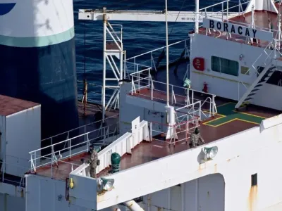 An aerial picture which shows a large tanker ship. It is white and burgundy. There are two masked soldiers in camouflage on the ship which is in the middle of the ocean. The name of the ship, Boracay, is clearly marked on it. 
