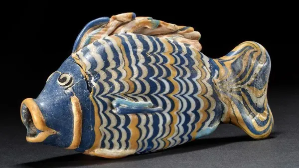 A blue, yellow and white glass vessel in the shape of a fish. Its body, tail and top fin have wavy stripes in blue, yellow and white, its face is blue, its open mouth outlined in yellow. It is resting on a black surface.