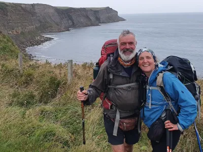 Zoe and Mike smiling with their arms around each other near a cliff overlooking a sea. They are both wearing shorts and waterproof coats, with bags on their back.