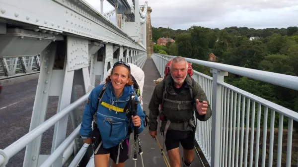 Zoe, wearing a blue waterproof coat and shorts, and Mike, in a green waterproof coat and shorts, hiking and smiling on Clifton Suspension Bridge. Zoe has sunglasses on her head and Mike has grey short hair.