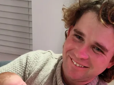 James Nunan has long curly brown hair and is smiling while cradling a baby in his arms. He is sitting on a sofa in a living room.