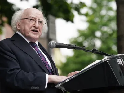 President of Ireland Michael D Higgins speaks at a lectern. We wears a black suit with a white shirt and and a purple floral tie. 