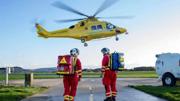 Yellow helicopter about to land, with two air ambulance workers in uniform walking to towards it